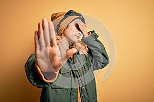 Beautiful asian skier girl wearing snow sportswear using ski goggles over yellow background covering eyes with hands and doing