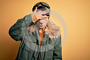 Beautiful asian skier girl wearing snow sportswear using ski goggles over yellow background peeking in shock covering face and
