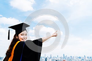 Beautiful asian university or college graduate student woman in graduation academic dress or gown, smiling and pointing at copy sp