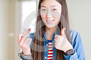 Beautiful Asian woman applying red lipstick on lips happy with big smile doing ok sign, thumb up with fingers, excellent sign