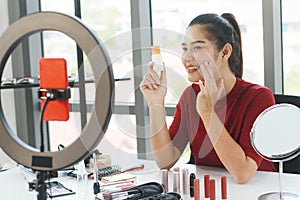 Beautiful Asian woman blogger hand holding Sunscreen bottle for skin care and recording on red smartphone for make up tutorial