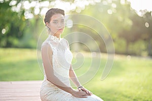 Beautiful asian woman bride in white wedding dress sit on wooden floor with green grass and tree in background