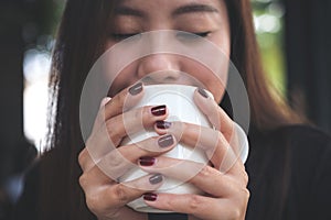 A beautiful Asian woman closing her eyes and holding a white cup of hot coffee with feeling happy in cafe