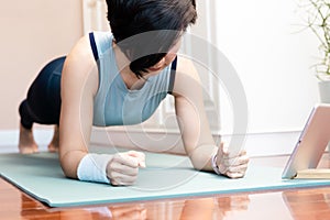 Beautiful Asian woman doing yoga plank pose to strengthening her core body and build muscles with computer tablet in bedroom at