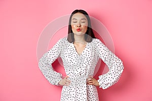 Beautiful asian woman in dress pucker lips and close eyes, waiting for kiss, standing over pink background