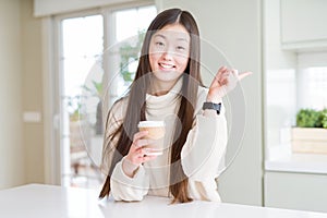 Beautiful Asian woman drinking a coffee in a take away paper cup very happy pointing with hand and finger to the side