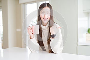 Beautiful Asian woman drinking a cup of coffee pointing and showing with thumb up to the side with happy face smiling