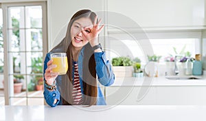 Beautiful Asian woman drinking a glass of fresh orange juice with happy face smiling doing ok sign with hand on eye looking