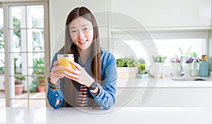 Beautiful Asian woman drinking a glass of fresh orange juice with a happy face standing and smiling with a confident smile showing