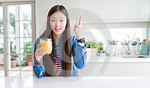 Beautiful Asian woman drinking a glass of fresh orange juice surprised with an idea or question pointing finger with happy face,