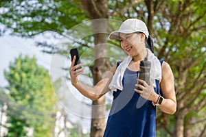 Beautiful Asian woman drinking water after jogging in the park. She is taking a picture of a water bottle with her smartphone