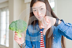 Beautiful Asian woman eating green fresh broccoli with angry face, negative sign showing dislike with thumbs down, rejection