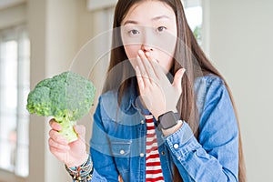 Beautiful Asian woman eating green fresh broccoli cover mouth with hand shocked with shame for mistake, expression of fear, scared