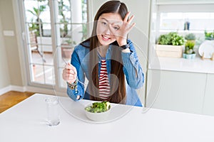 Beautiful Asian woman eating green fresh broccoli with happy face smiling doing ok sign with hand on eye looking through fingers