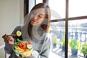 A beautiful asian woman eating and holding a bowl of fresh mixed vegetables salad