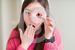 Beautiful Asian woman eating pink sugar donut cover mouth with hand shocked with shame for mistake, expression of fear, scared in