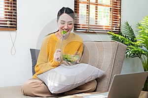 Beautiful Asian woman eats salad with a wide smile, happy smiling face and surfs the internet with laptop computer