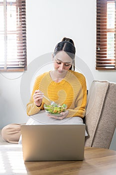 Beautiful Asian woman eats salad with a wide smile, happy smiling face and surfs the internet with laptop computer