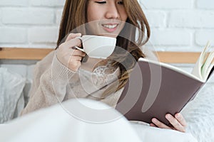 Beautiful asian woman enjoying warm coffee and reading book on bed in her bedroom.