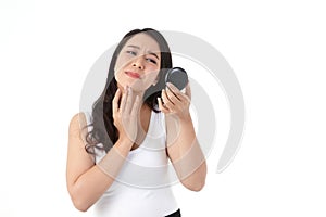 A beautiful Asian woman feels stressed. She is holding a mirror, looking at acne on her cheeks.