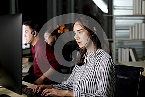 Beautiful Asian woman with headphones work night shift at call center customer care service desk, looking at desktop computer,