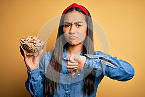 Beautiful asian woman holding bowl with healthy corn flakes cereals over yellow background with angry face, negative sign showing