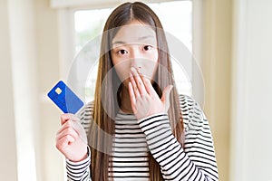 Beautiful Asian woman holding credit card cover mouth with hand shocked with shame for mistake, expression of fear, scared in