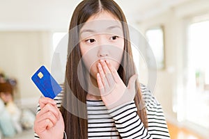 Beautiful Asian woman holding credit card cover mouth with hand shocked with shame for mistake, expression of fear, scared in