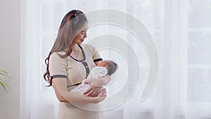 Beautiful Asian woman holding newborn baby in her arms standing in front of windows at cozy home.Happy infant baby sleep in mother