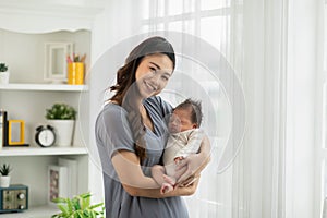 Beautiful Asian woman holding newborn baby in her arms standing in front of windows at cozy home.Happy infant baby sleep in mother