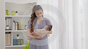Beautiful Asian woman holding newborn baby in her arms standing in front of windows at cozy home.Happy infant baby sleep in mother