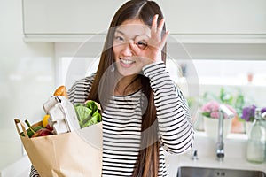 Beautiful Asian woman holding paper bag of fresh groceries with happy face smiling doing ok sign with hand on eye looking through