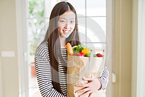 Beautiful Asian woman holding paper bag of fresh groceries with a happy face standing and smiling with a confident smile showing