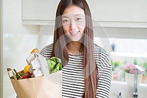 Beautiful Asian woman holding paper bag of fresh groceries with a happy face standing and smiling with a confident smile showing