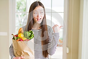 Beautiful Asian woman holding paper bag of fresh groceries pointing and showing with thumb up to the side with happy face smiling