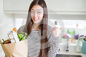 Beautiful Asian woman holding paper bag of fresh groceries pointing and showing with thumb up to the side with happy face smiling