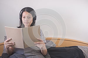 Beautiful Asian woman listening music with headphone and reading book relaxing on the bed.