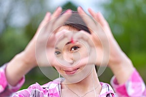 Beautiful asian woman looking through heart gesture made with hands in summer green park