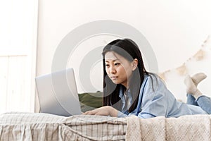 A beautiful Asian woman is lying on the bed and working using a laptop from home. A young woman works at home in