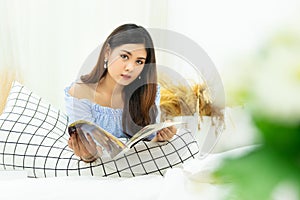 Beautiful Asian woman lying on white cozy bed reading a book enjoys of rest at comfort home