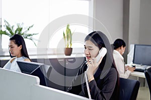 Beautiful Asian woman operator officer talking with customer by using landline phone at working desk in office with busy working