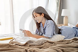 Beautiful Asian woman in pajamas is using her phone, replying to messages on her bed in her bedroom