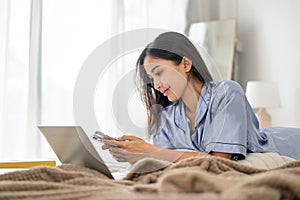 A beautiful Asian woman in pajamas is using her phone, replying to messages while working on her bed