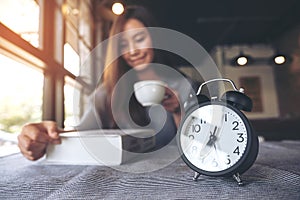 Asian woman reading book and drinking coffee in the morning with black alarm clock on table