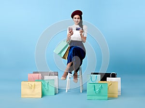 Beautiful Asian woman shopper sitting and carrying shopping bags with using credit card and mobile phone in hands on blue