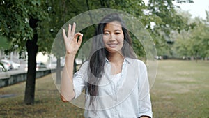 Beautiful Asian woman showing OK hand gesture outside in city park smiling alone