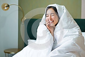 Beautiful asian woman sitting on bed, covered in blankets and duvet, laughing and smiling, enjoying weekend in bedroom