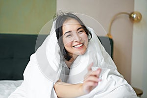 Beautiful asian woman sitting on bed, covered with white duvet, smiling, enjoying happy weekend morning, laughing at