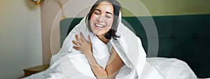 Beautiful asian woman sitting on bed, covered with white duvet, smiling, enjoying happy weekend morning, laughing at