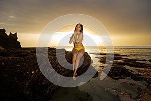 Beautiful Asian woman sitting on the rock, wearing yellow bikini. Slim body. Sunset on the beach. Summer vacation. Bright sunlight